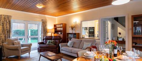 A cozy living room with a wooden ceiling, a fireplace, a dining area set for a meal, and a bookshelf.