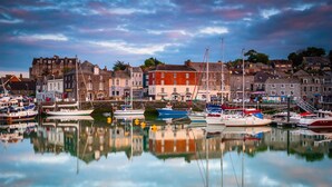 A harbor with boats docked, buildings in the background, and a calm water surface reflecting the structures. - Cottage in Cornwall near Sandy Beach (Rock)