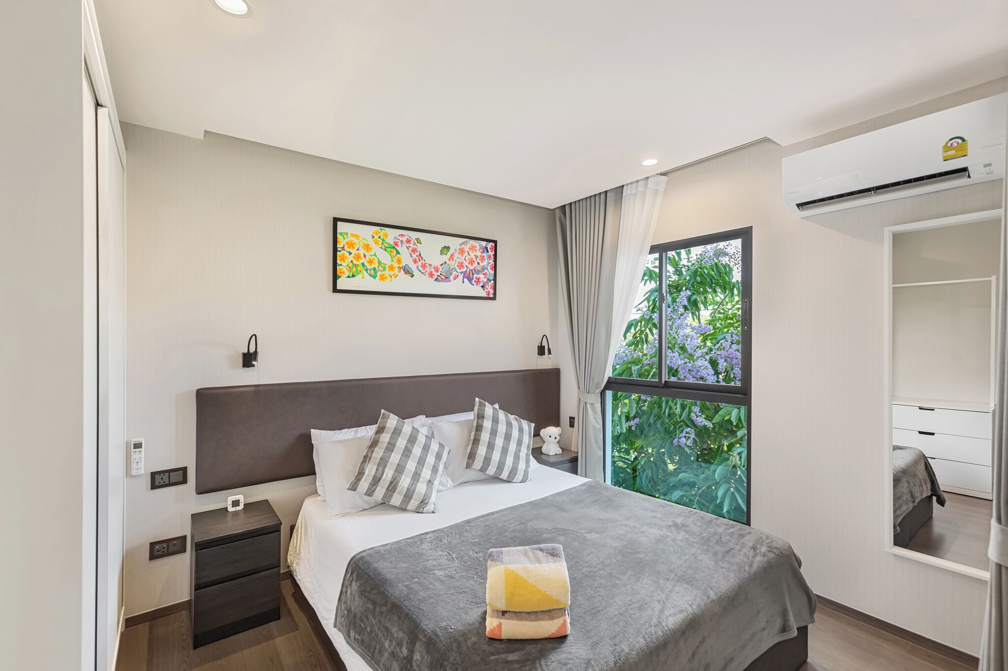 A modern hotel room with a bed, bedside tables, a window with a view of greenery, and a wall-mounted air conditioning unit.