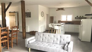 A modern living room with a grey sofa, wooden chairs, a small round stool, and a kitchen area with white cabinets and a white refrigerator.