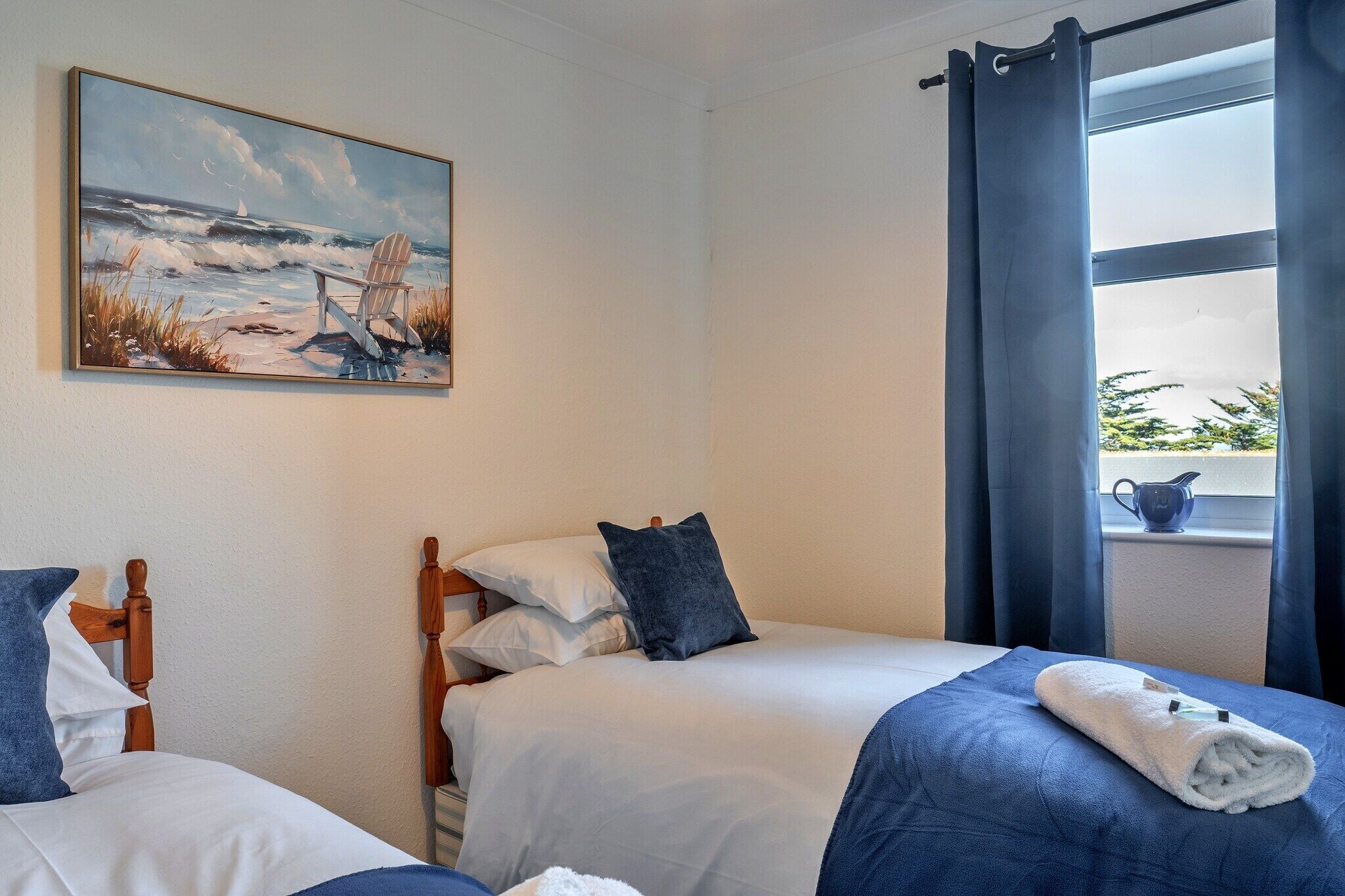 A bedroom with two beds, a painting of a beach scene, a window with curtains, and a view of greenery outside.