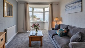 A living room with a grey sofa, a wooden coffee table with a vase of flowers, a radiator, and a view of the sea through a bay window. - Apartment in St Ives near Porthminster Beach (St Ives)