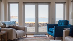 Un soggiorno con un divano color beige, una poltrona blu e un tappeto bianco e soffice. Grandi vetrate conducono a un muro in pietra e offrono vista sul mare.