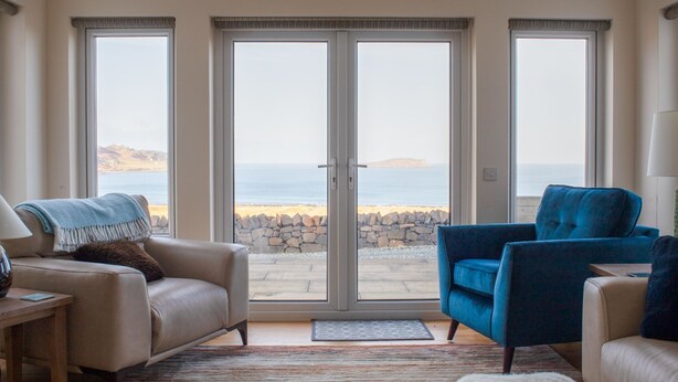 A living room with a beige sofa, a blue armchair, and a white furry rug. There are large glass doors leading to a stone wall and a view of the sea. - solas Alainn by Interhome (North Skye)