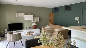Een moderne woonkamer met een flatscreen televisie, een bank, een eettafel en een bijzettafeltje met een lamp.