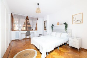 A bedroom with a bed, bedside table, chair, and a window with sheer curtains.