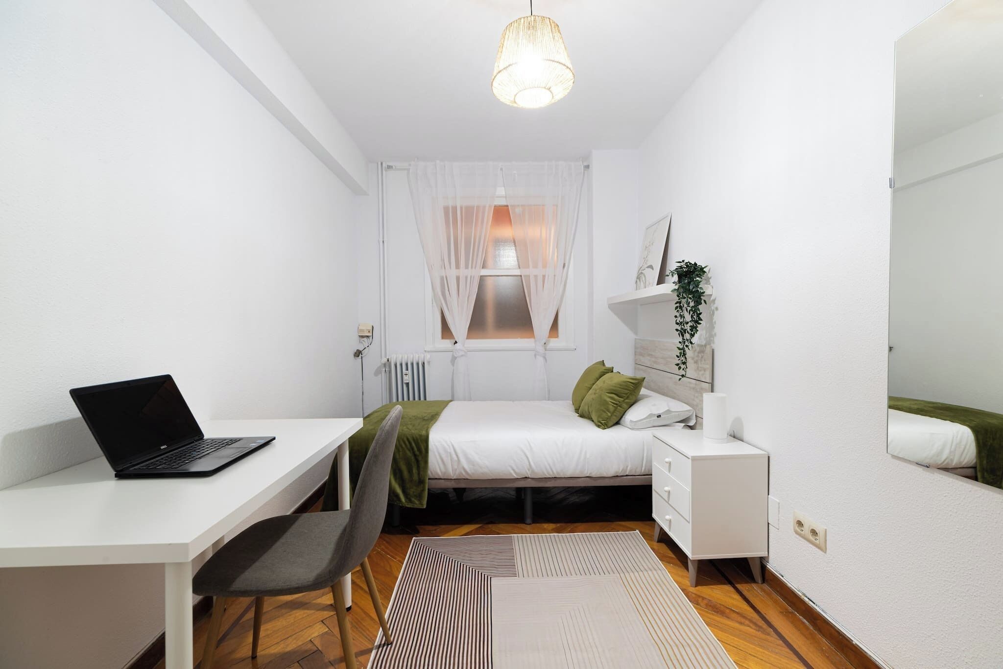 Une chambre à coucher comprenant un lit, un bureau avec un ordinateur portable, une chaise, une table de chevet, un miroir et un radiateur.