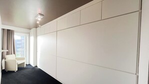 A hotel room with a large white panel wall, a small white chair, a desk, and a window with curtains.