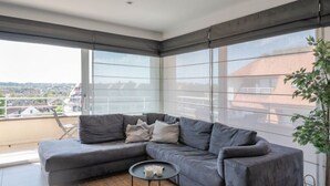 Un salon moderne avec un canapé d’angle gris, une petite table basse ronde et une grande fenêtre donnant sur la ville.