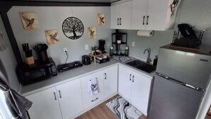 A compact kitchen with white cabinets, a stainless steel refrigerator, a microwave, a coffee maker, and a sink. There are wall decorations including a tree and birds, and a patterned rug on the floor.