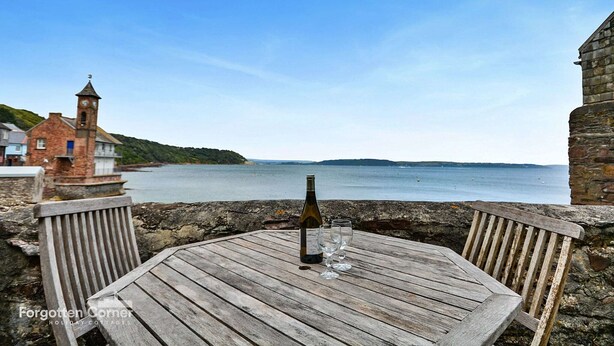A wooden table with a bottle and glasses, overlooking a calm sea and a distant island. - Cottage in Kingsand by Stunning Beach Views (Cawsand)