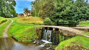 Un pont de pierre rustique enjambe un petit ruisseau, entouré d’une végétation luxuriante, avec un bâtiment historique en arrière-plan.