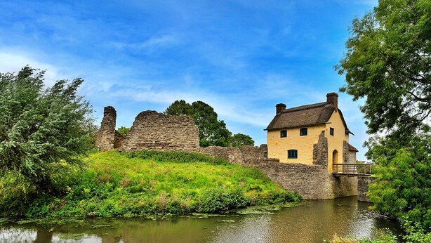 Un bâtiment historique en pierre, doté d’une façade jaune et d’un toit de chaume, situé au bord d’une rivière paisible.