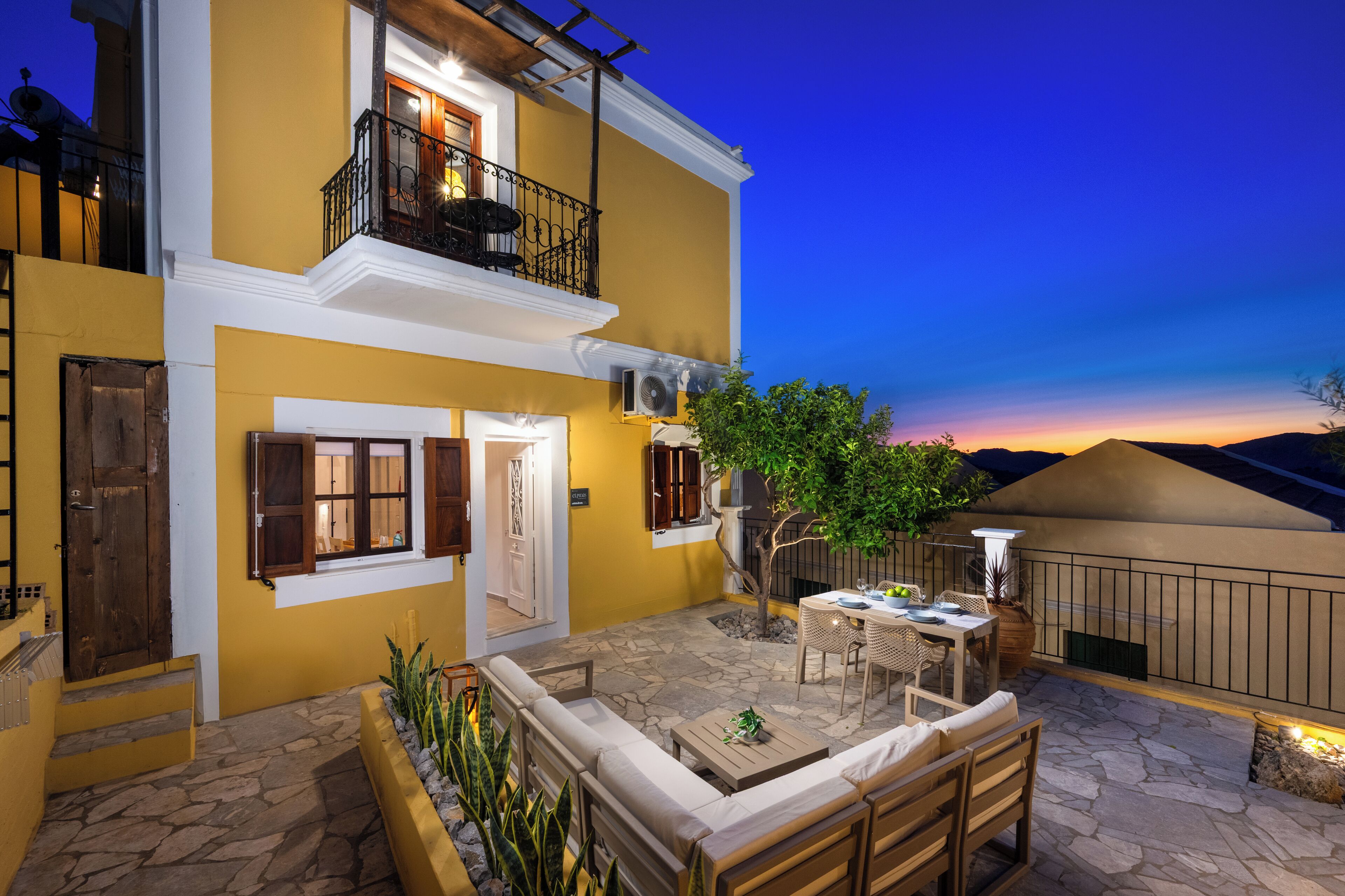 A two-story building with a yellow facade, a balcony with black railings, and a stone patio area with outdoor furniture.
