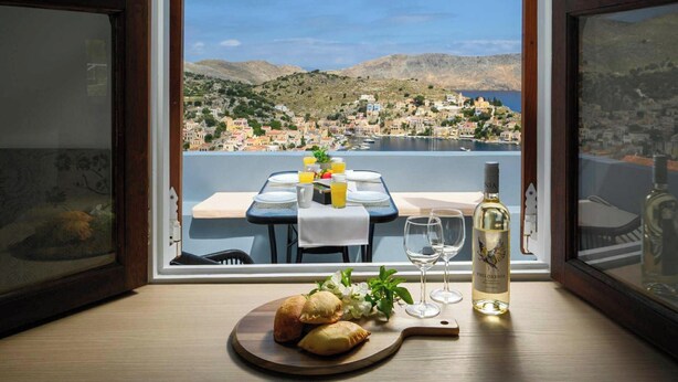 Une table à manger avec une bouteille de vin Philokemia, un verre d’eau et une assiette de nourriture, le tout placé devant un paysage pittoresque d’une ville côtière.