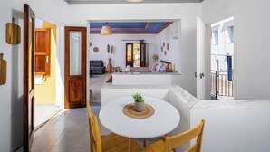 Un coin repas avec une table ronde, des chaises et une plante en pot. On aperçoit un salon avec un canapé et une fenêtre.