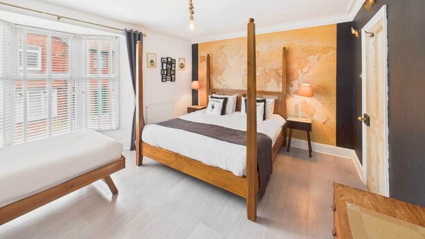Un dormitorio con cama con dosel, una mesita de noche, una lámpara, un mapa mundial en la pared y una ventana con persianas.