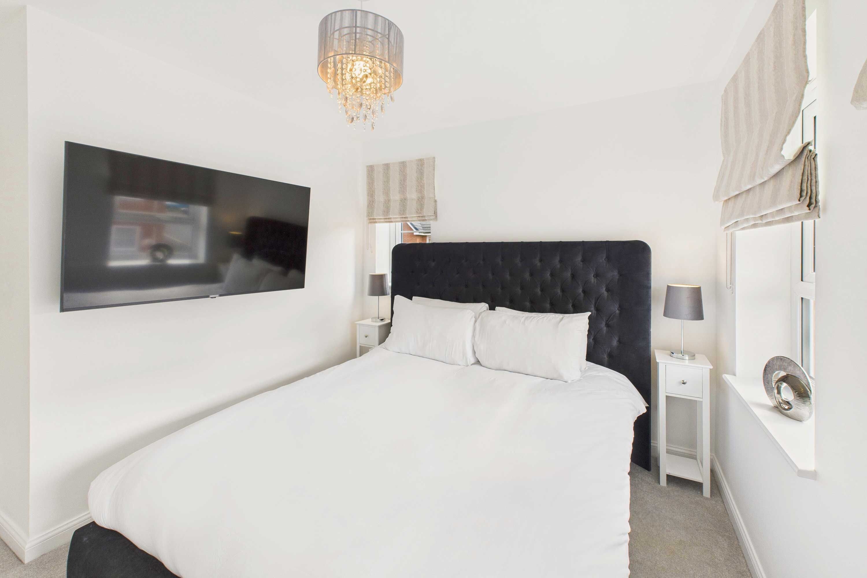 A modern bedroom with a large bed, a flat-screen TV mounted on the wall, a bedside table with a lamp, and a decorative mirror.
