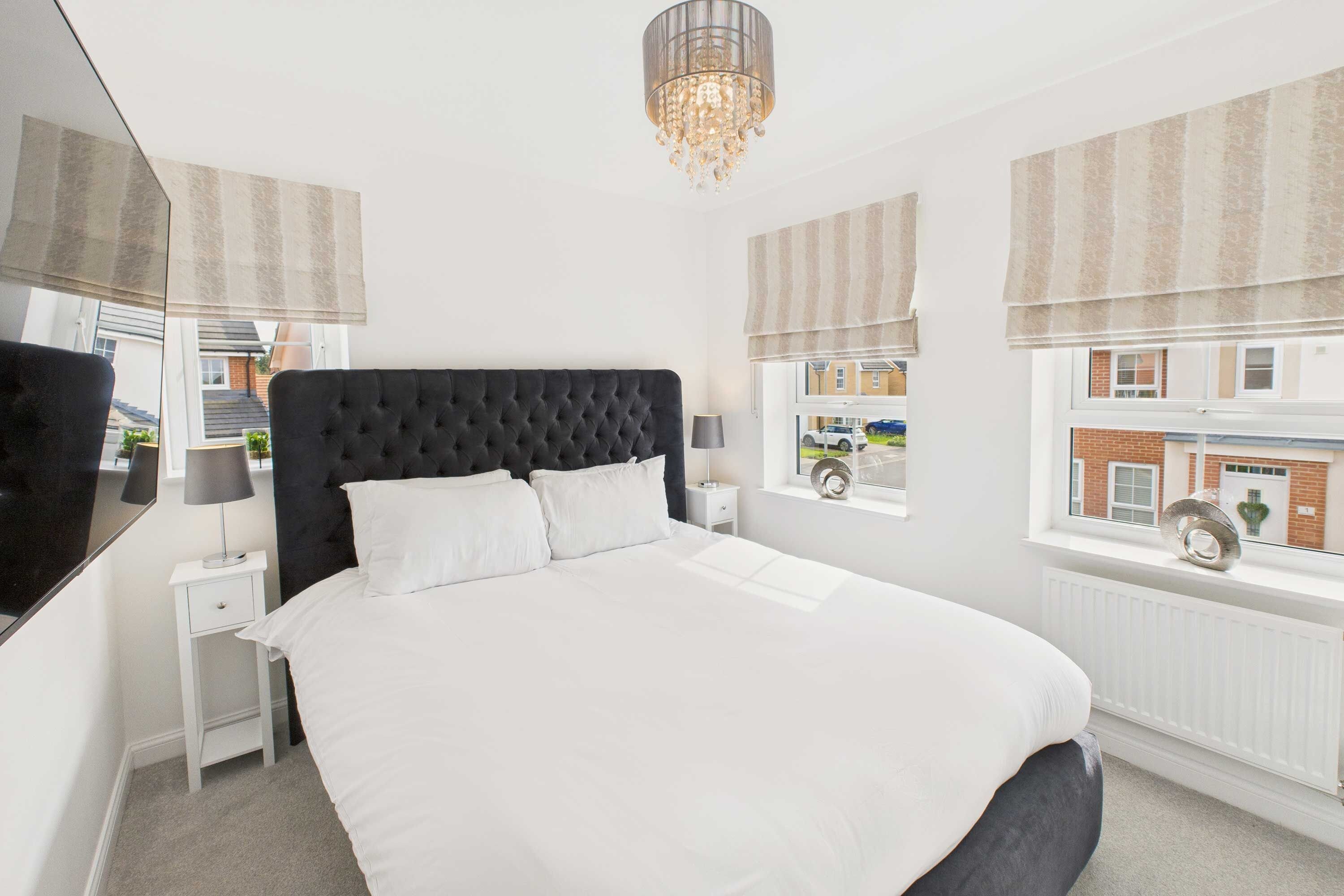 A bedroom with a large bed, a bedside table, a lamp, a chandelier, and windows with blinds.