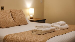 A neatly made bed with a beige patterned bedspread and pillows, a stack of white towels, and a bedside table with a lamp. - 2 bedroom cottage for 4 guests, No Deposit (Dorrington)