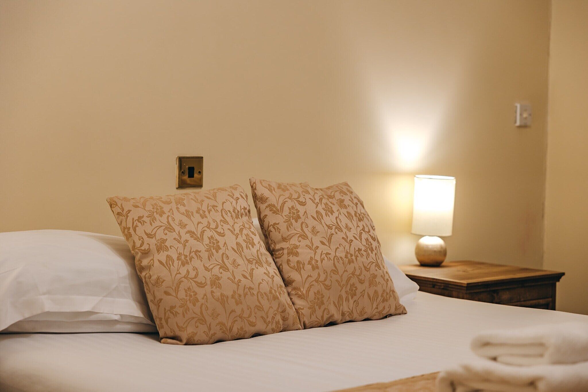 Een net opgemaakt bed met bloemenkussens en een bedlampje.