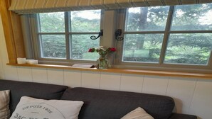A room with a window overlooking a forest, a shelf with a vase of flowers, and a sofa with decorative pillows.