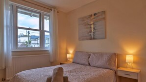 A bedroom with a large bed, two bedside lamps, a window with curtains, a radiator, and a painting on the wall.