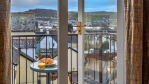 A balcony with a table holding pastries and a glass of orange juice, overlooking a town with houses and a hill in the distance.