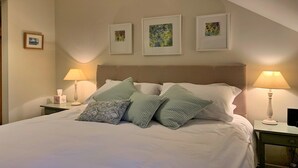Ein ordentlich bezogenes Bett mit weißen Leinen und gemusterten Kissen, zwei Nachttische mit Lampen und drei gerahmte Kunstwerke an der Wand.