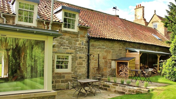 Ein Steinhaus mit Ziegeldach, einem großen Fenster und einer Terrasse mit Tisch und Stühlen.