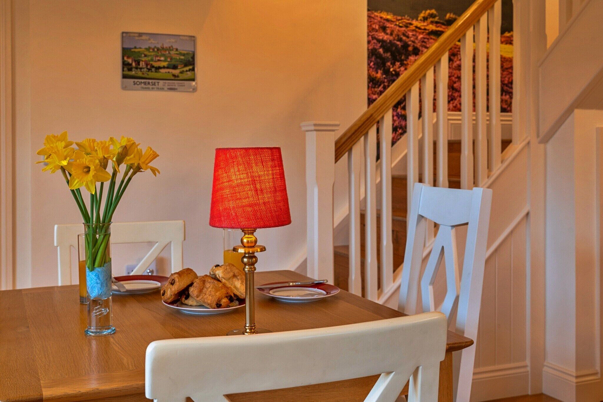 A dining table with a vase of daffodils, a red lamp, and plates with food.