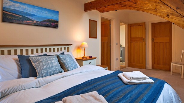 A bedroom with a large bed, a wooden ceiling beam, a painting of a landscape, and a view of a door and another room. - Cottage in Minehead near Exmoor National Park (Minehead)
