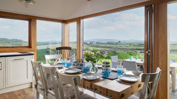 A dining area with a table set for four, large windows offering a view of the countryside, and a kitchen with white cabinets.