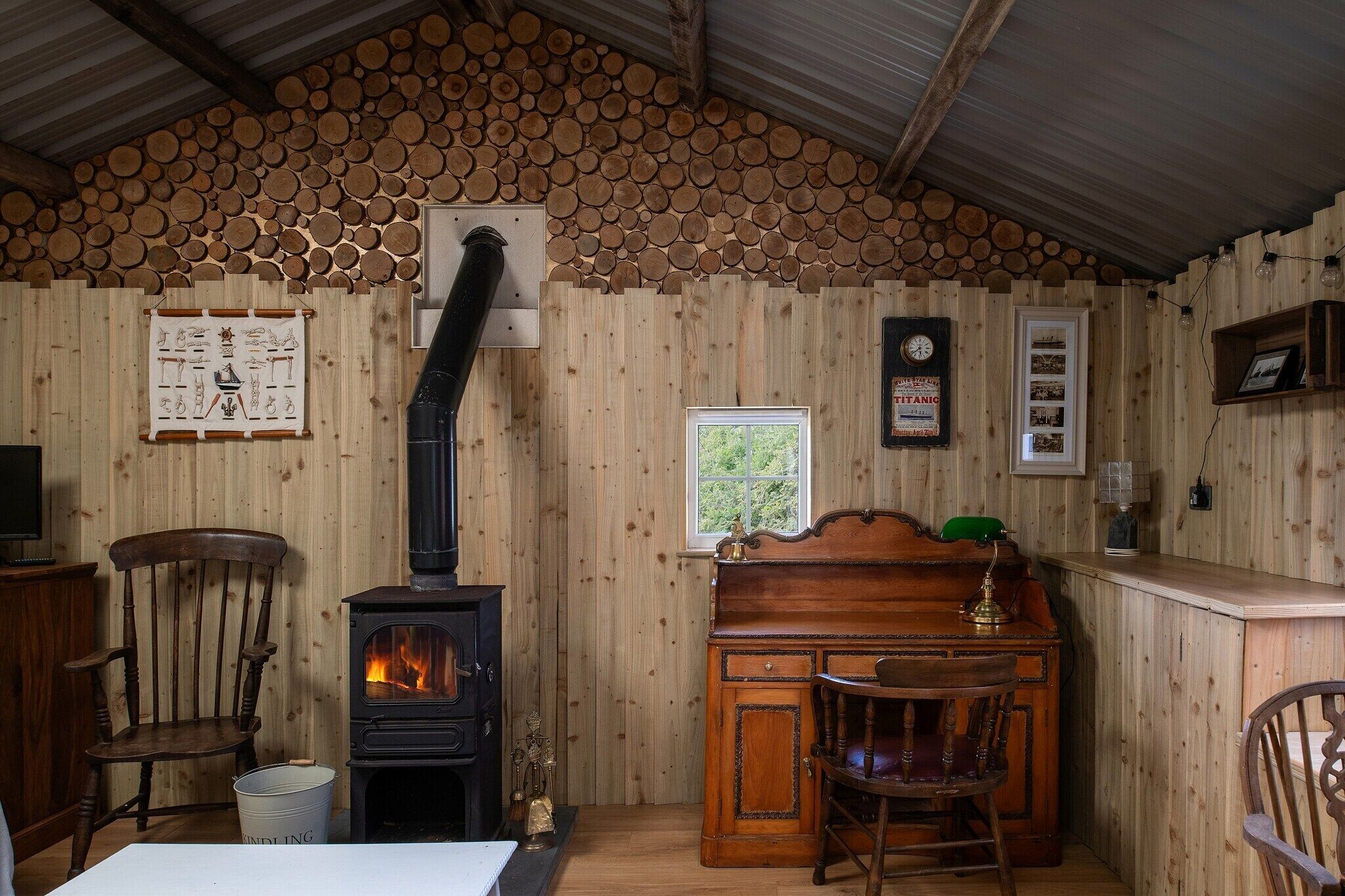A rustic cabin interior with a wood-burning stove, wooden walls, a wooden desk, and a chair.