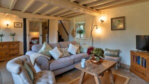 A cozy living room with a sofa, a coffee table, and a television. There is a staircase leading to another room, and a wooden cabinet with books. - Cottage in Dorset near Lulworth Cove (Winterborne Houghton)
