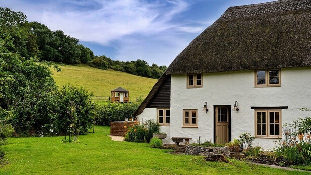A white house with a thatched roof, surrounded by a lush green garden and a hill in the background. - Cottage in Dorset near Lulworth Cove (Winterborne Houghton)