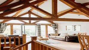 A spacious living area with a high ceiling and exposed wooden beams, a sofa set, a dining table, and large windows offering a view of the outdoors. - Barn in Wales with Swim Spa & Scenic Views (Aberystwyth)