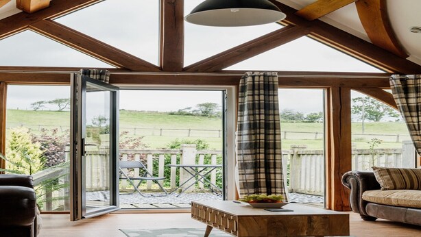 A spacious room with a large sliding glass door leading to an outdoor patio. The room features a wooden ceiling, a black pendant light, a brown leather sofa, and a wooden coffee table with a bowl of fruit. - Barn in Wales with Swim Spa & Scenic Views (Aberystwyth)