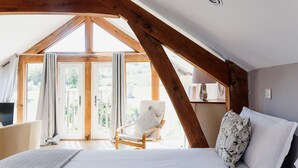 A bedroom with a sloped ceiling, wooden beams, a bed, a chair, and a view through large windows. - Barn in Wales with Swim Spa & Scenic Views (Aberystwyth)