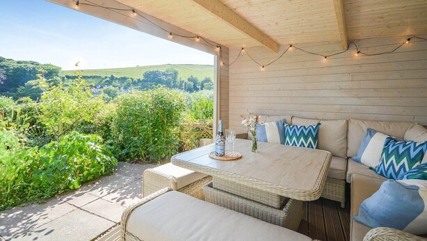 An outdoor patio with wicker furniture, a table with a bottle and glasses, and string lights. - Holiday Home in Kingsand near the Beach (Kingsand)