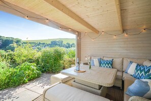 An outdoor patio with wicker furniture, a table with a bottle and glasses, and string lights. - Holiday Home in Kingsand near the Beach (Kingsand)