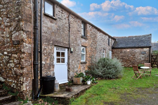 A stone cottage with a white door, a small garden with a wooden table and chairs, and a sign reading "Sam Cottage".