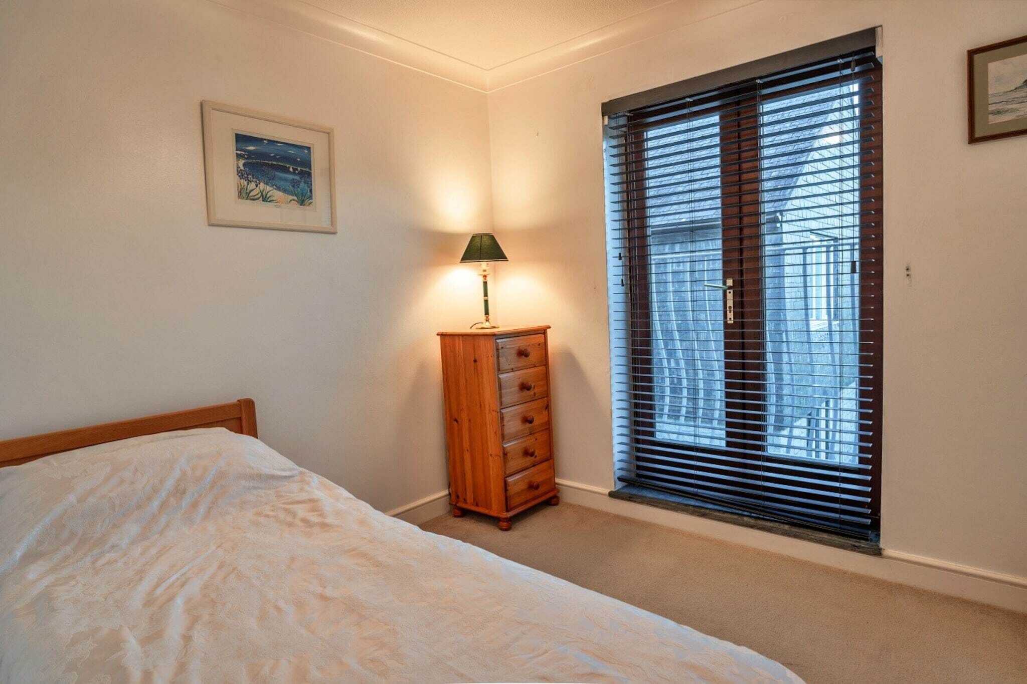 A bedroom with a bed, a wooden dresser, a nightstand with a lamp, a window with blinds, and a framed picture on the wall.