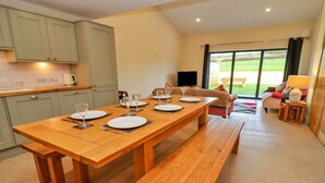 A dining area with a wooden table set for four, a sofa, and a view of a garden through a glass door. - Cottage in Cotswolds with Hot Tub & Garden (Little Rissington)
