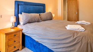 A bedroom with a blue headboard, a wooden nightstand with drawers, a lamp, and a door. - Barn in Cotswolds near Bourton-on-the-Water (Little Rissington)