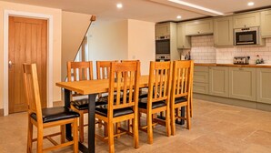 A dining area with a wooden table and chairs, a kitchen with green cabinets and a microwave, and a wooden door. - Barn in Cotswolds near Bourton-on-the-Water (Little Rissington)