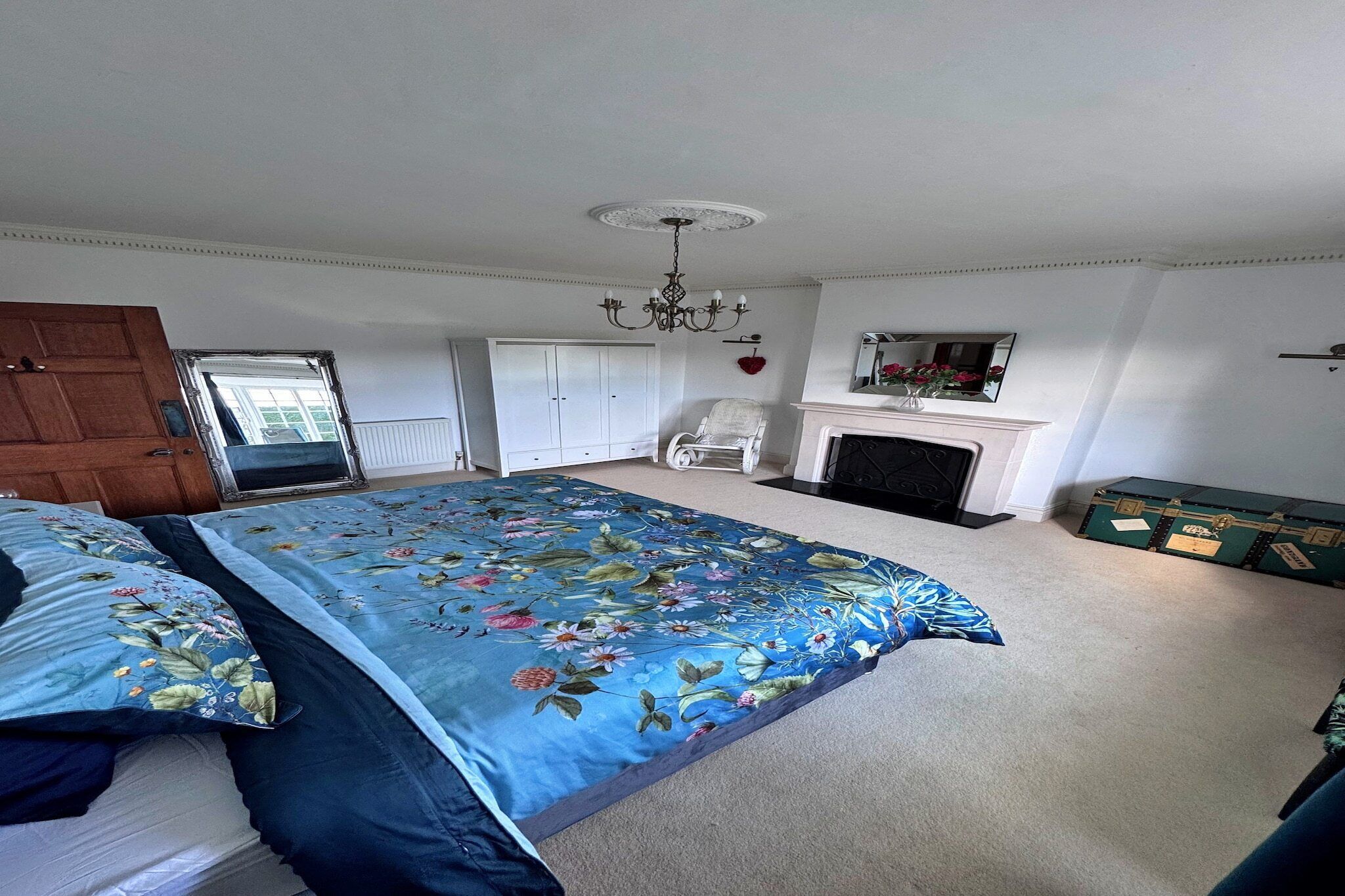 A bedroom with a large floral bedspread, a white chair, a fireplace, and a green trunk.