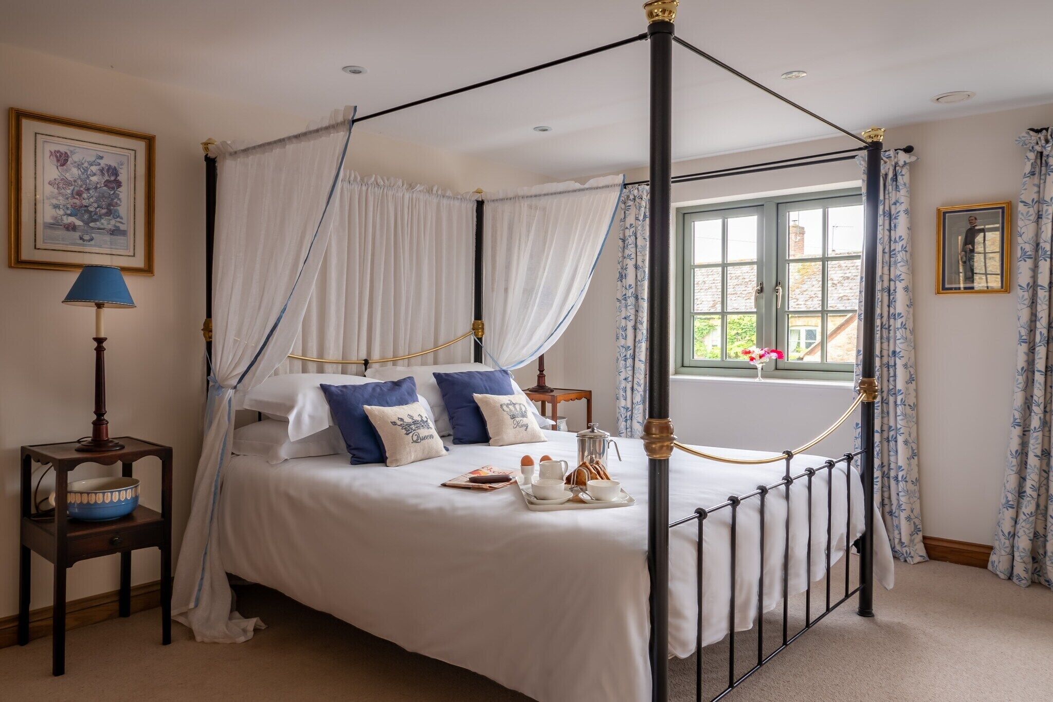 A four-poster bed with white bedding and blue pillows, a bedside table with a lamp, and a window with curtains.