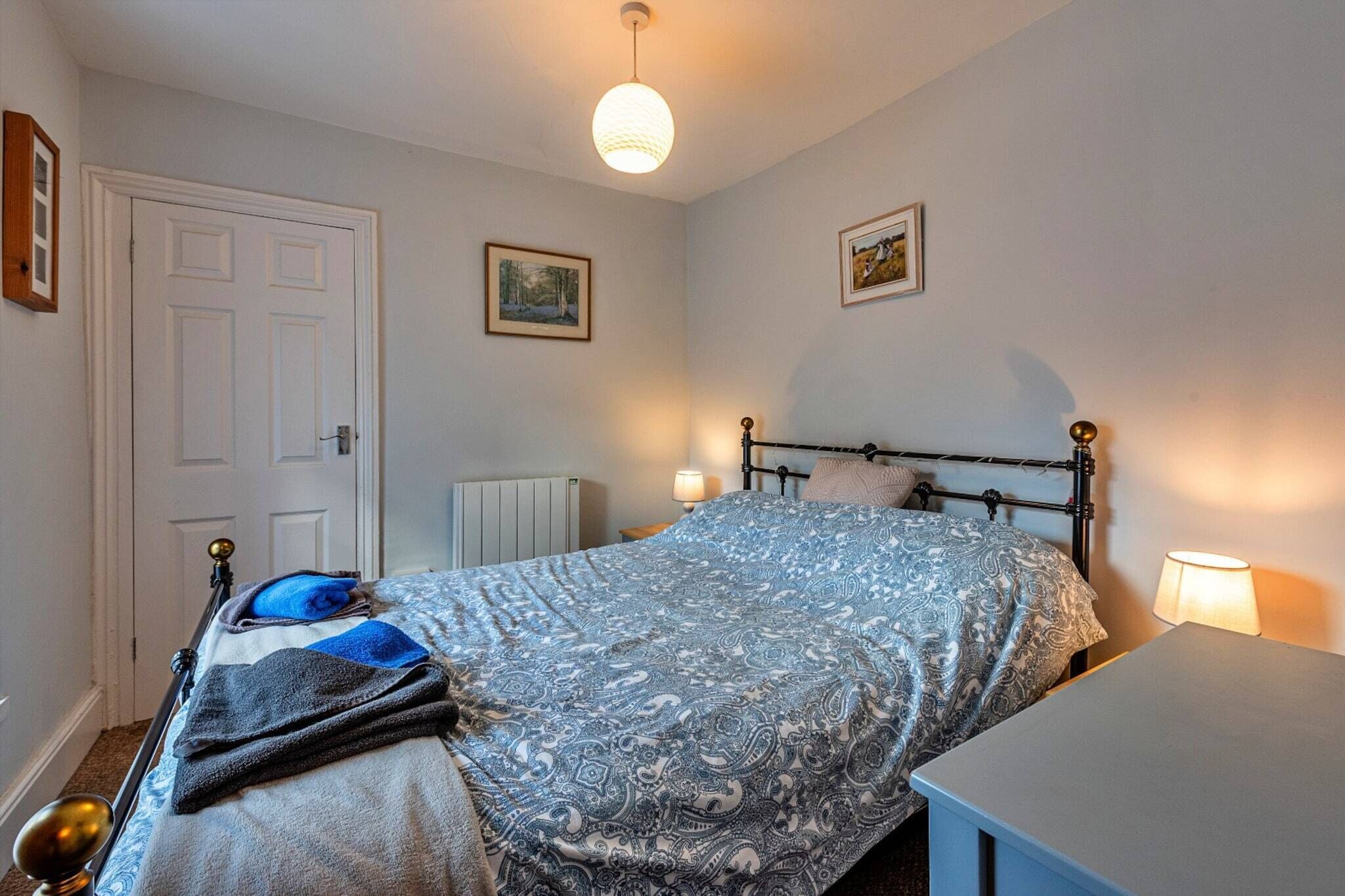 A bedroom with a bed, bedside tables, a radiator, and two framed pictures on the wall.