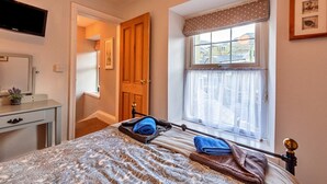Un dormitorio con cama, mesita de noche, televisor montado en la pared, ventana con cortinas y puerta que da a otra habitación.